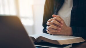 Women's hands are folded in prayer on a Holy Bible over a laptop at work