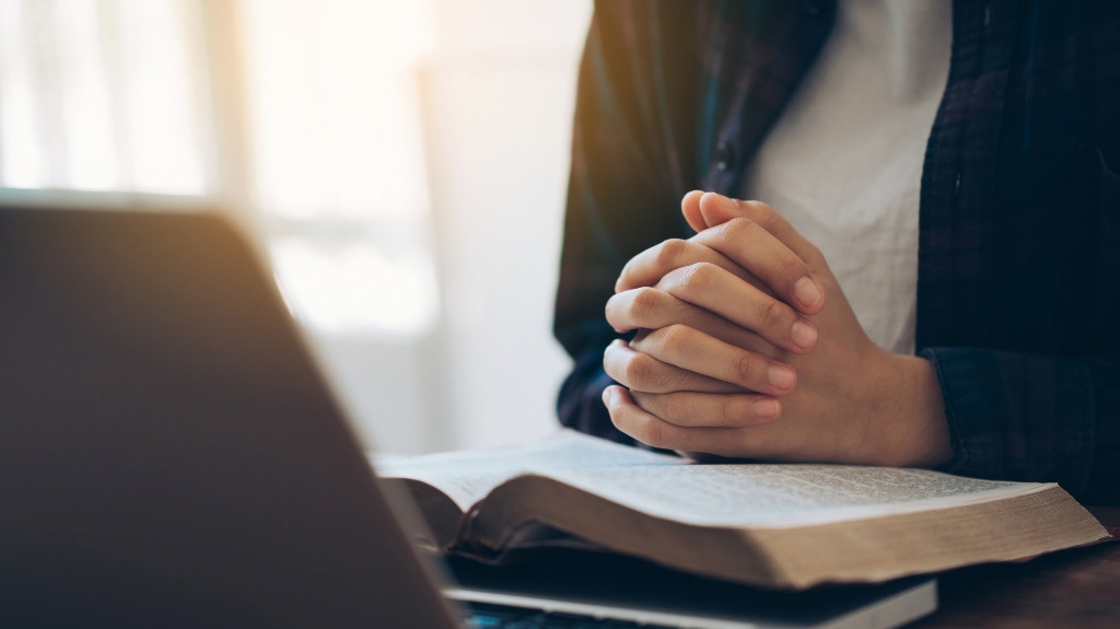 Women's hands are folded in prayer on a Holy Bible over a laptop at work