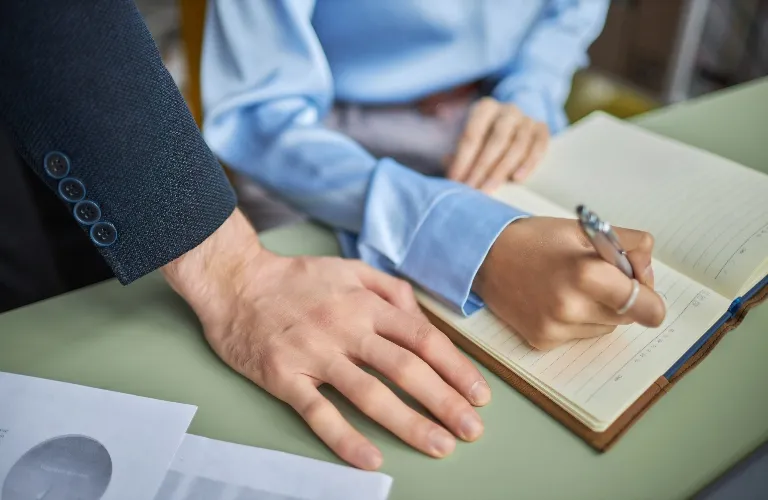 Two individuals in professional attire engaged in writing and reviewing documents on a desk.