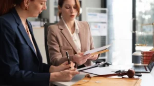 Two professional women reviewing documents at a white desk with legal books, a gavel, and a laptop.