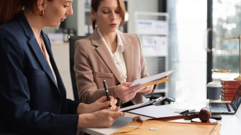Two professional women reviewing documents at a white desk with legal books, a gavel, and a laptop.