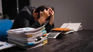 Man in black suit sitting at desk with large stack of documents and laptop, holding head in hands.