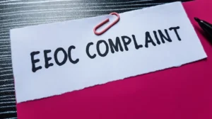 White paper with "EEOC COMPLAINT" text clipped by pink paperclip on red folder, black pen nearby.