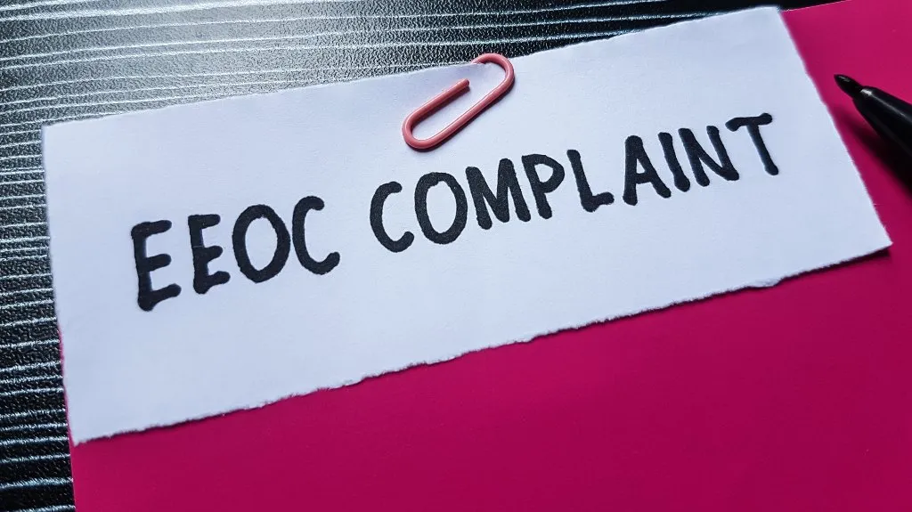 White paper with "EEOC COMPLAINT" text clipped by pink paperclip on red folder, black pen nearby.