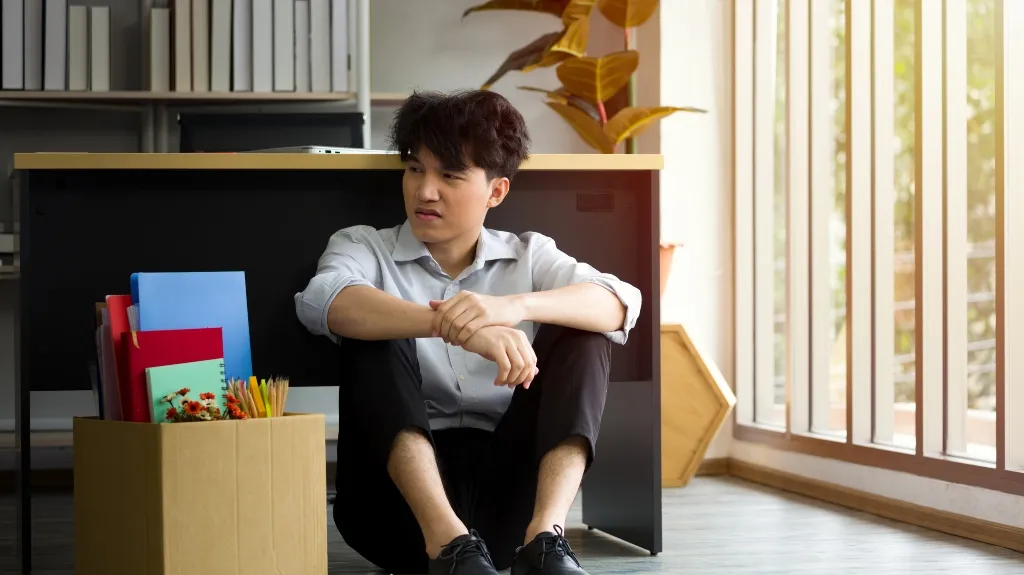 Young man in light blue shirt sitting on office floor next to cardboard box with files and stationery.