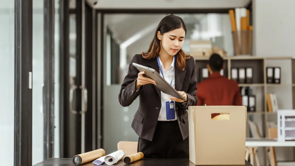 Professional woman in black suit packing documents into a cardboard box in office.