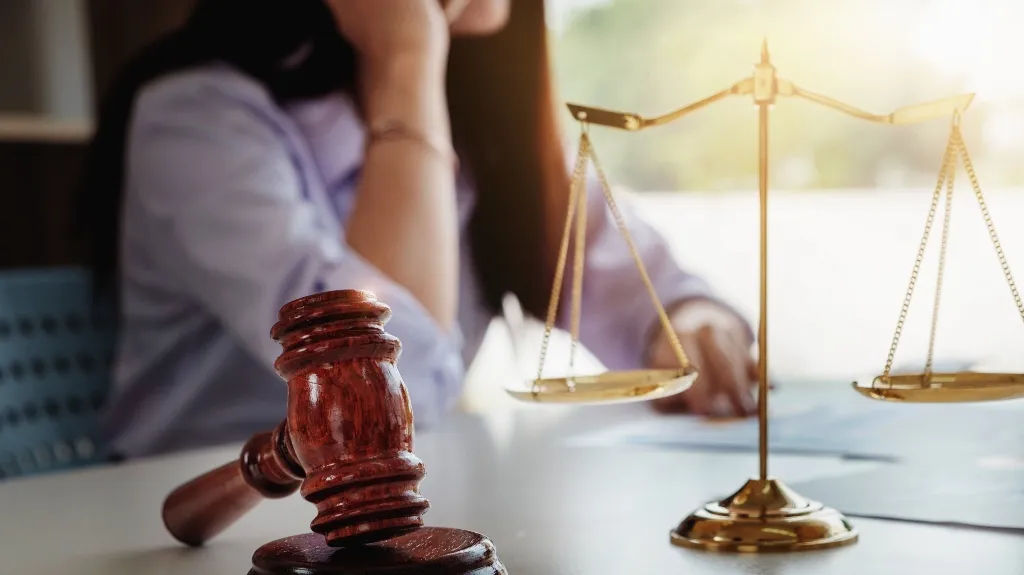Wooden judge's gavel and golden scales of justice on a desk with blurred person in background.