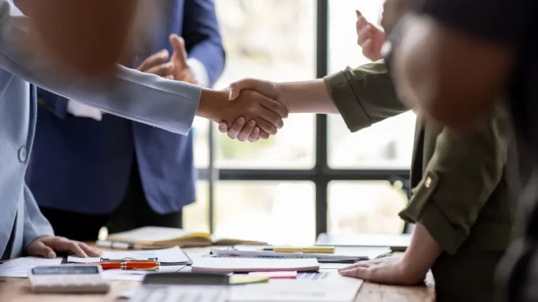Two business professionals shaking hands over a desk with documents and office supplies.