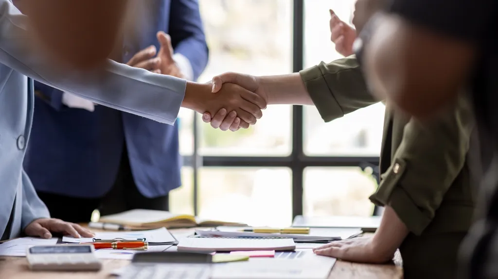 Two business professionals shaking hands over a desk with documents and office supplies.