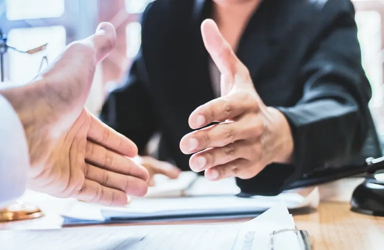 Two people in formal attire reaching out to shake hands over a desk with documents.