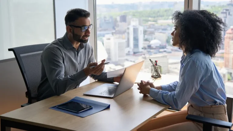 Two professionals engaged in a discussion at an office desk with a laptop and tablet.