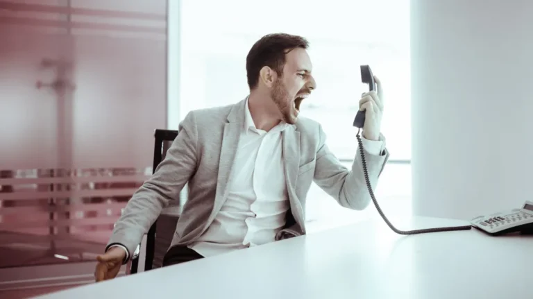 Man in gray blazer angrily shouting into a black office telephone handset at a white desk.
