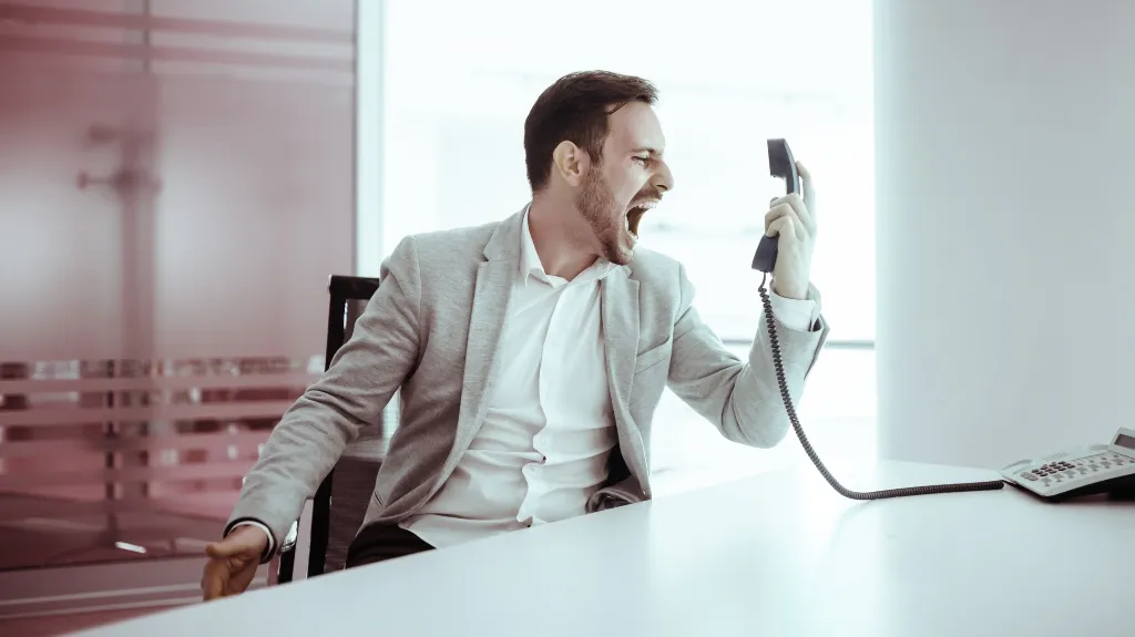 Man in gray blazer angrily shouting into a black office telephone handset at a white desk.